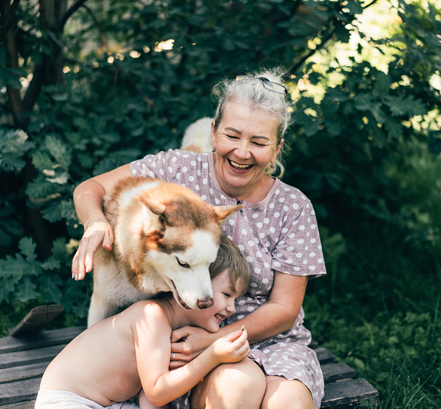 A grandmother hugs her son and husky dog on a bench outdoors.