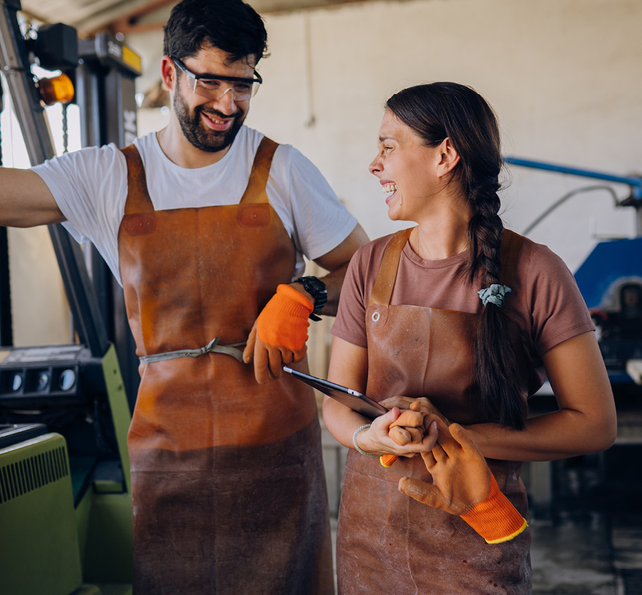 A man and woman in leather aprons laughing together in a factory.